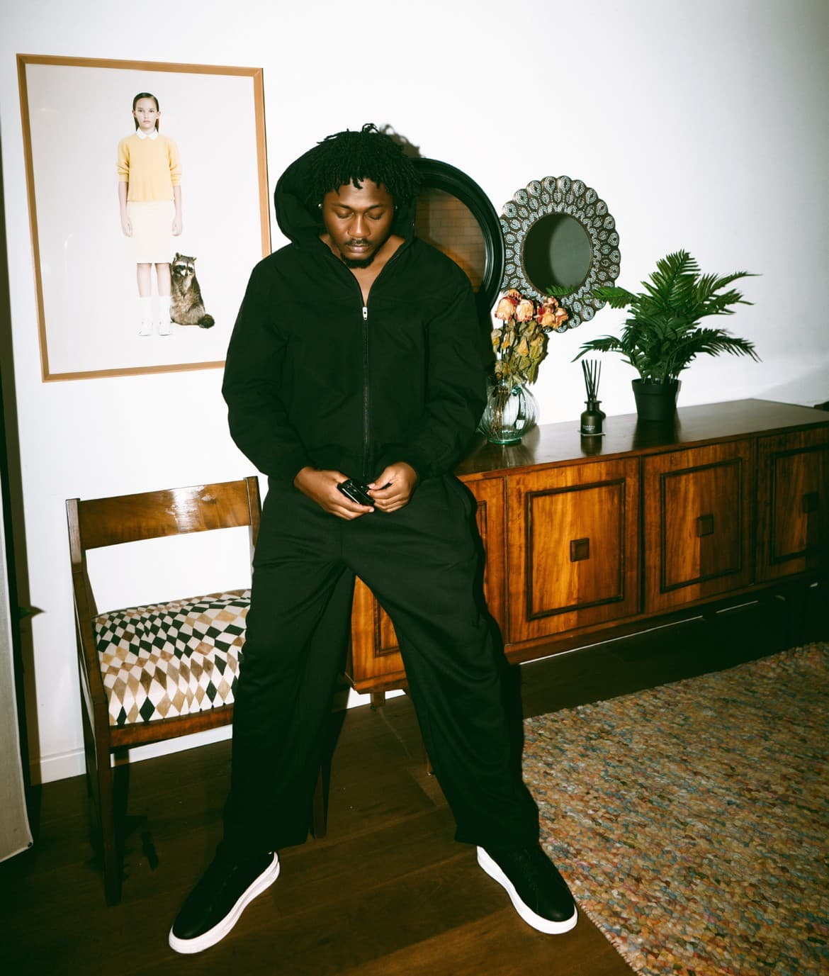 Phantom in a black jacket and trousers, leaning against a wooden sideboard at home, looking down